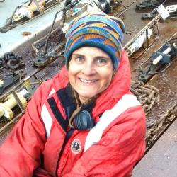 Woman with blue and orange beanie wearing a red coat smiling with ship deck in background