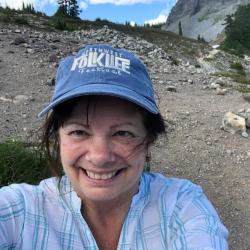 Brunette woman with blue hat and white shirt smiling with hills in background.
