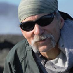 Man with bandana, glasses and mustache with a green coat, water and mountains in background. 