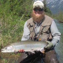 Man with hat, glasses and beard holding a fish with water and mountains in the background