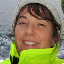 Brunette woman wearing white hat and yellow coat with ocean in background. 