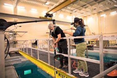 Scientists: Jay Barlow and Shannon Rankin of SWFSC MMTD are leaning over the railing of a walkway that spans the horizontal length of the Ocean Technology Development Tank inside SWFSC. Jay Barlow is holding a drifting buoy and lowering it into the tank for equipment testing. 