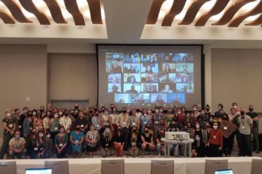2022 DCLDE Group photo of in person participants in the foreground and virtual participants in the background projected onto a screen. 