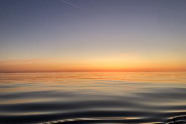 A color image taken at sunrise from the deck of a research ship. The water is calm and the rising sun’s light brightens the horizon at the image’s center.