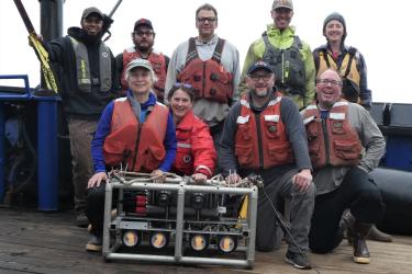 A group of scientists pose around an underwater drop camera in a metal housing aboard a ship. Credit: NOAA Fisheries