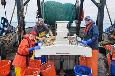 hree people standing on deck of a trawl fishing vessel posing for a picture as they sort catch on a sorting table.