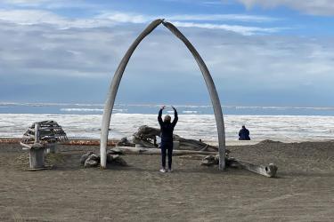 Mabel Baldwin-Schaeffer stands under an arch made of two whale ribs.  She has her arms raised, with her back to the camera and facing the Arctic Ocean on a beach in Utqiagvik, Alaska. The skeleton of a small overturned boat is propped up on the beach beside the arch. Sea ice gathers along the shore and in the distant offshore with a stretch of open water in between. Blue sky and gray clouds are visible above the horizon.