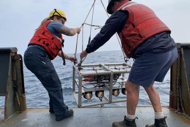 photo of two people in hard hats and orange vests lowering stereocamera array overboard 