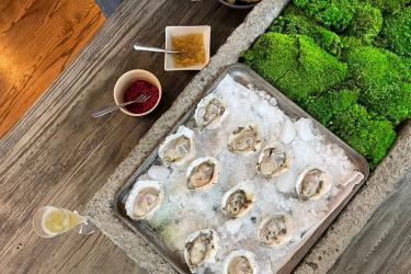 A raw bar with oysters on ice, next to a glass of champagne and two mignonette sauce options for the oysters.