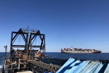 A color image taken on a sunny day from the deck of a commercial clam vessel. The lower half of the image shows the deck, the twin dredges mounted to the stern of the fishing vessel, and the conveyor belt that moves the catch from the dredges to the ship. The container ship is in the middle right side of the image, stacked with containers, sitting on the horizon. The word “cosco” is painted in large letters on its side.
