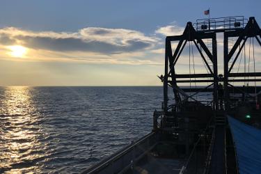 A color image taken on an early morning. The right half of the image is the back deck of a commercial clam vessel, twin dredges rise to the top of the image. At left, the sun is masked by a few clouds, rising over the horizon on a calm sea.