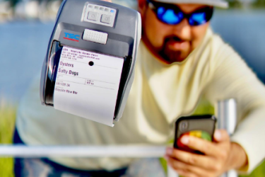 A shellfish grower holding a BlueTrace printer and oyster label in one hand, and a smartphone in the other hand.