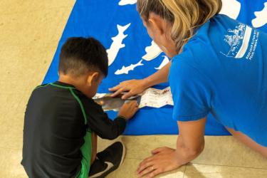 NOAA staff members sits next to a child to help them learn how to measure a pretend fish using the Hawaii nearshore fisheries regulations guide.