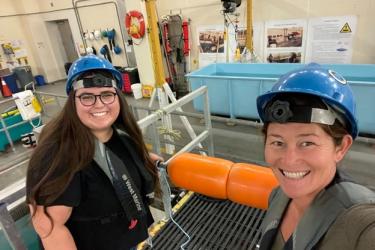 Kourtney Burger and Cory Hom-Weaver stand on a walkway that spans across the technology tank at Southwest fisheries science center. They wear hard hats and dawn life jackets for safety. The are testing out a surface buoy configuration. 