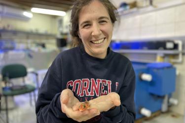 A woman in a dark blue sweater holds a small orange fish in her open palms.