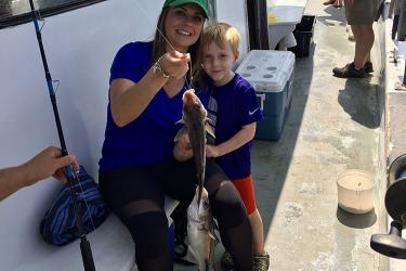 A white woman wearing a green hat and blue shirt sits on a boat next to a boy. She is holding a fish they caught.