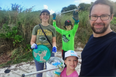 Heloise is on the beach with her son, daughter, and husband. They are holding trash collectors, buckets, and have gloves on after doing a beach clean up.