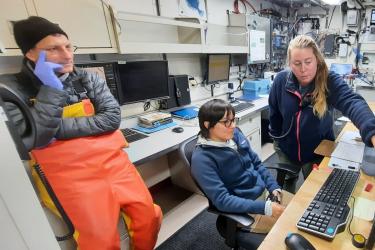  A female scientist sits in front of a computer with a male scientist standing to the left of her and a female scientist standing to the right. The standing female scientist points to something on the computer screen. 