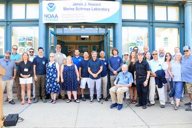 A large group of scientists stands in front of the open glass doors of a building. A banner above them reads: “James J. Howard Marine Science Laboratory” and “NOAA Fisheries”.