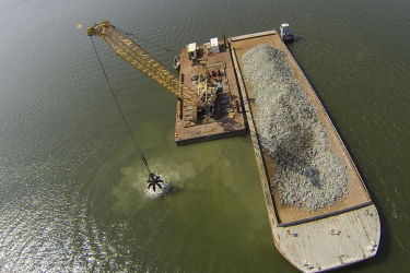 An aerial view of a crane moving hard substrate from the deck of a barge into the Piankatank River