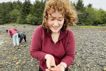 Virginia Pearson stands on a rocky beach in Sitka, Alaska holding a small purple shore crab in their hands.