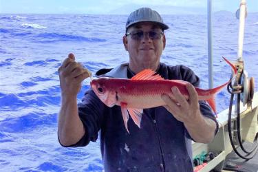 A fisher smiles while holding up a red and white fish, called an onaga, with both hands while out at sea.