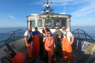 Seven scientists stand in front of a research vessel’s wheelhouse near the bow while at sea. Some are wearing orange foul weather pants, baseball hats, t-shirts, and sunglasses. The sea is calm and there is a light layer of fog in the background hovering above the water. The sky is blue and there are a few white, wispy clouds. 