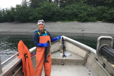 Teacher at Sea and Commercial Fisherwoman, Germaine Thomas, stands at the bow of her boat. She is dressed in orange coveralls, cold weather gear, and is holding two salmon--one in each hand.