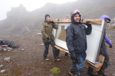 Four people bundled up against the cold are carrying a new door across the terrain. The door is vertical and the people are carrying it using wooden rods propped on their shoulders. Rocky hills and fog are in the background.