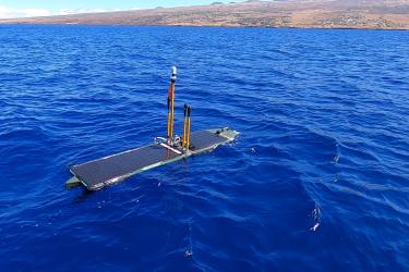 A rectangular autonomous surface vehicle floats on the ocean’s surface off the coast of a Hawai‘i shoreline.