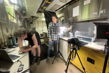 Two scientists stand in a lab taking photos of fish in a tank. There is a camera and lighting system set up. One scientist is adjusting the lighting on one side of the lab while the other scientist is on the other side of the lab in front of a laptop. Both scientists are looking at the laptop screen.
