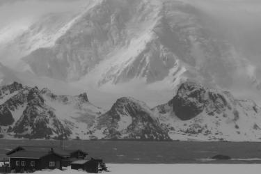 Black and white photo of the old Cape Shirreff field camp sitting in snow in the foreground. Behind that is a strip of ocean, and behind that are snow-covered mountains with the tops shrouded in clouds.