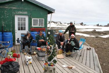 The Cape Shirreff team celebrates Christmas outside. In the left top corner is a green camp building with a red window cover leaning against the wall and four blue water storage barrels pushed up against the wall. There is a white door with nine small windows, and a white window to the right of the door. In the foreground are wooden decking, several camping chairs, a trash bag with wrapping paper, some plastic bins and cardboard boxes that are serving as tables, and some plates, mugs, and gifts. In the cent