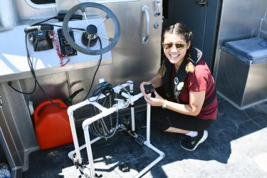 Onboard a boat, a young woman wearing sunglasses and holding a small camera crouches near a frame made from PVC pipe.