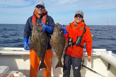 Two people stand on the back of a boat next to each other posing for the camera. Each is holding a similarly sized flatfish