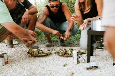 Four people squat on a gravel driveway eating plates of clams cooked on a propane burner.