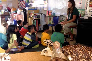 The Honu Project coordinator Lauren Kurpita sits on a chair in front of young students seated on the floor in a classroom as she conducts sea turtle outreach at Naʻālehu Elementary School.