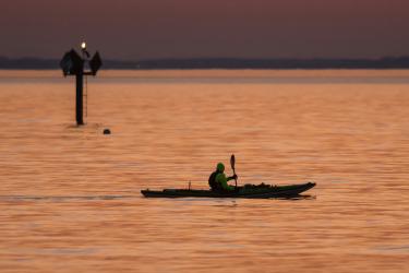 A person in a kayak paddles by a day marker at sunrise