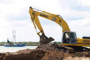 An excavator digs into the soil alongside a river, with a fishing boat in the background