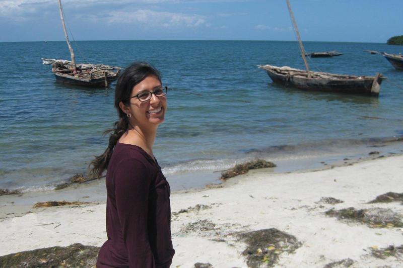 Nicole Naar standing on a tropical beach in Tanzania, with small fishing boats floating on the water close by.