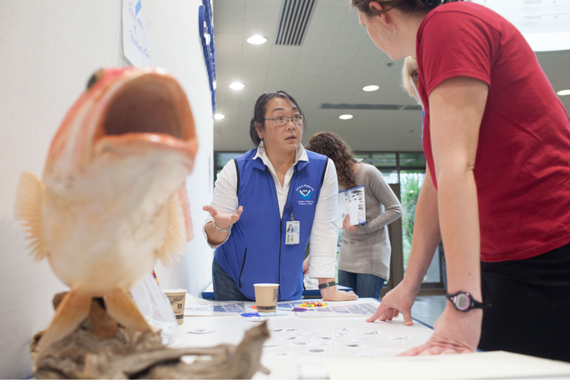 Photo of Lisa Hiruki-Raring talking with a visitor at a NOAA Fisheries information table.