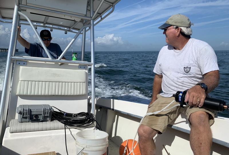 Two men are aboard a center-console boat. One drives, while the other holds scientific equipment.