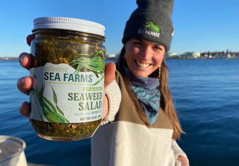 A smiling Atlantic Sea Farms employee stands on a dock while holding up a jar of 'Atlantic Sea Farms Fermented Seaweed Salad.'