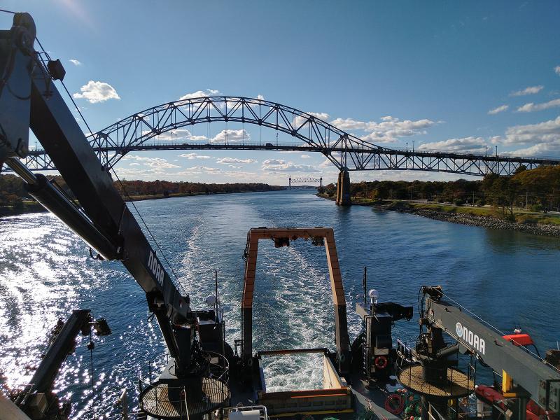  Color image taken during daylight from the upper deck of a research vessel, facing toward the rear of the vessel as it sails through a canal. Land is visible on each side of the canal, and the ship leaves a wake in the water. Two steel framed bridges are in the image. The ship has just passed under the bridge used by vehicles. The railroad bridge is in the distance. 