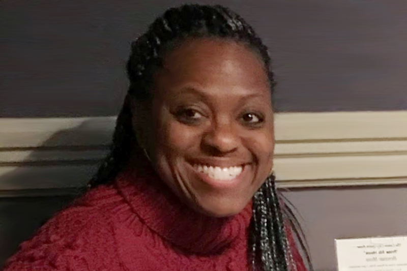 Medium close up of a black woman with long braids in a pink turtleneck sweater smiling for the camera.