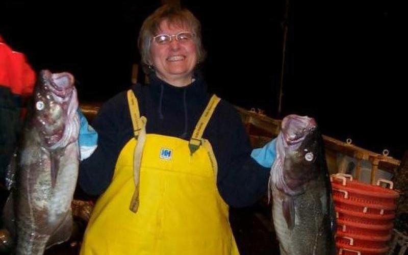 Women in foul weather gear overalls holds up two cod. In the background are baskets and fish on the deck for sorting from a bottom trawl