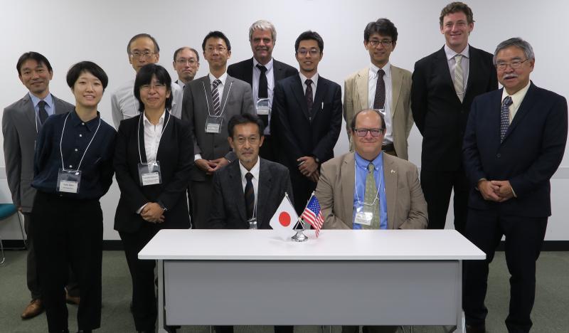 Co-chairs Hideaki Aono and Mike Rust are seated at a table with miniature Japanese and American flags; they are surrounded by 11 members of the UJNR panel.