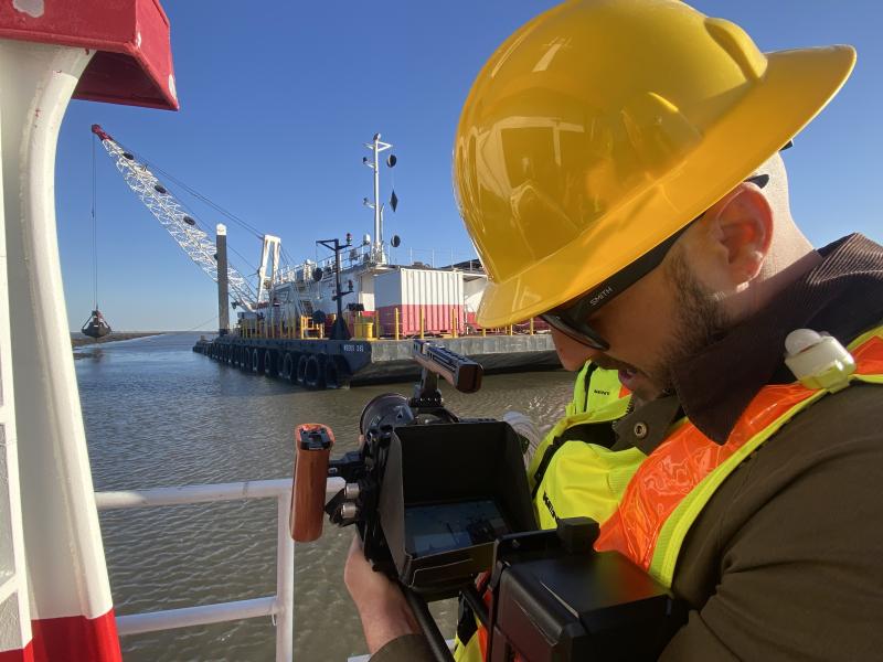 A man wearing a safety vest and hard hat takes a photograph of a barge