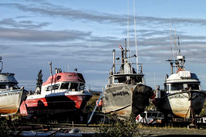 Photo of 4 small fishing boats lined up on dry land in the sun. 3 boats are gray with rust; the second from the left is red. 