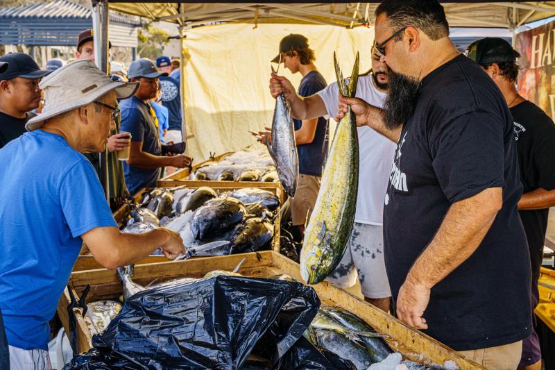 Customers point to whole fish on ice while fishmongers hold up a tuna and a mahi at an outdoor seafood market.